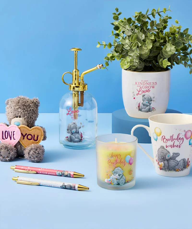 A Tatty Teddy themed gift display arranged on blue pedestals against a blue background, featuring a plush teddy bear holding two heart shapes reading ‘Love’ and ‘You,’ a gold‑top plant mister bottle printed with ‘Let love grow,’ a white plant pot reading ‘Plant kindness and grow love’ with a leafy green plant, a glass candle labelled ‘Happy Birthday,’ a white mug printed ‘Birthday wishes,’ and two matching pens placed in front. A Tatty Teddy themed gift display arranged on blue pedestals against a blue background, featuring a plush teddy bear holding two heart shapes reading ‘Love’ and ‘You,’ a gold‑top plant mister bottle printed with ‘Let love grow,’ a white plant pot reading ‘Plant kindness and grow love’ with a leafy green plant, a glass candle labelled ‘Happy Birthday,’ a white mug printed ‘Birthday wishes,’ and two matching pens placed in front.