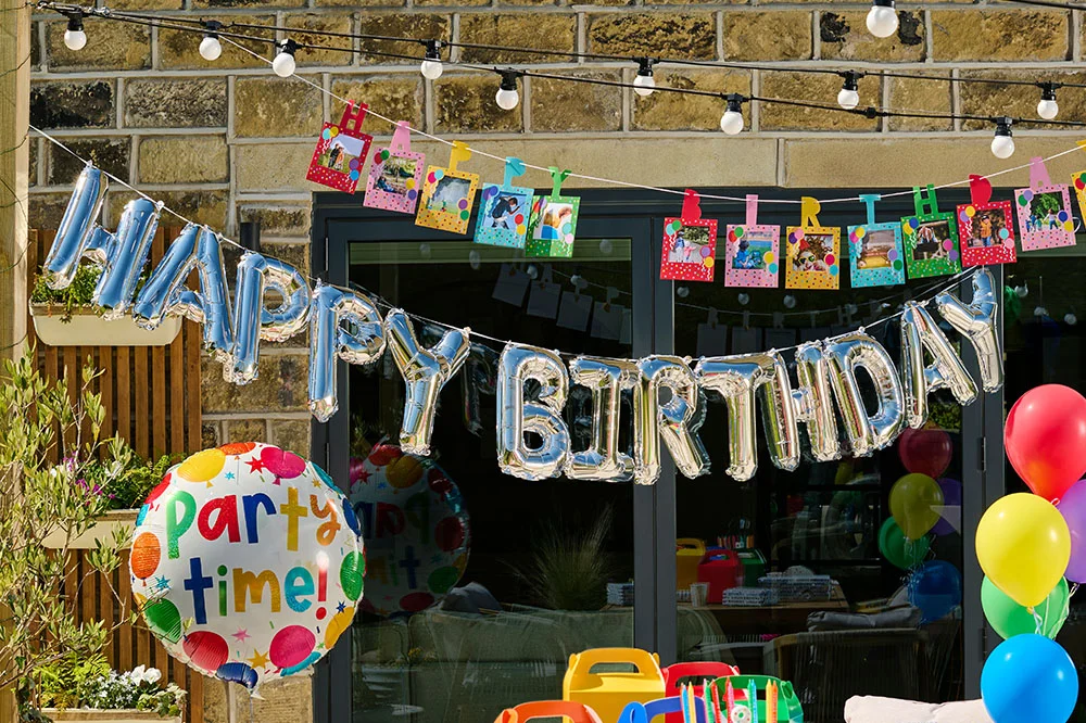 A birthday party display outside a house featuring a silver foil ‘Happy Birthday’ balloon banner, colourful photo bunting with clipped photographs, a large ‘Party Time!’ balloon, and clusters of rainbow-coloured balloons. Decorative honeycomb hanging ornaments and party decorations complete the vibrant celebration setup.