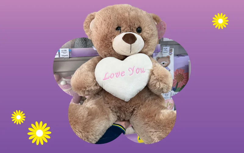 A soft brown teddy bear holding a white heart-shaped cushion embroidered with the words ‘Love You,’ displayed against a purple and pink gradient background with small yellow daisy illustrations.