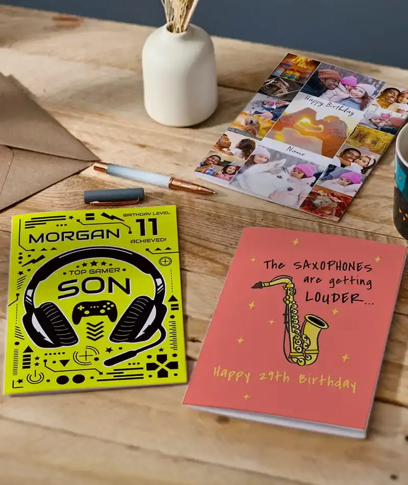 Selection of birthday cards, including personalised photo and gaming-themed designs, displayed on a wooden table with stationery accessories.