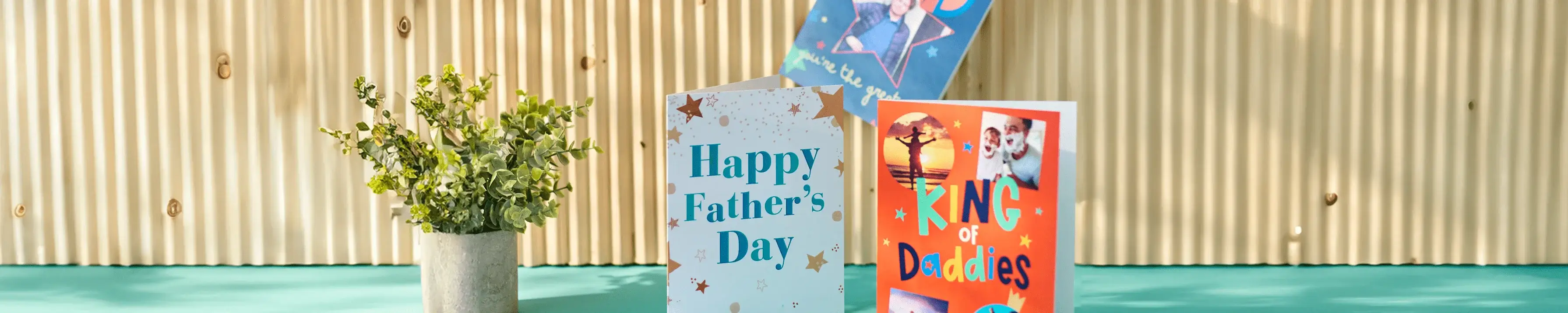 A Father's Day card with a beer mug illustration and the text 'HAPPY FATHER'S DAY!' is placed on a marble surface. Next to it is an open card with the message 'To Dad, wishing you a very Happy Father's Day. Love, [Name]'. The scene also includes a green candle, some books, and shadows of plants.