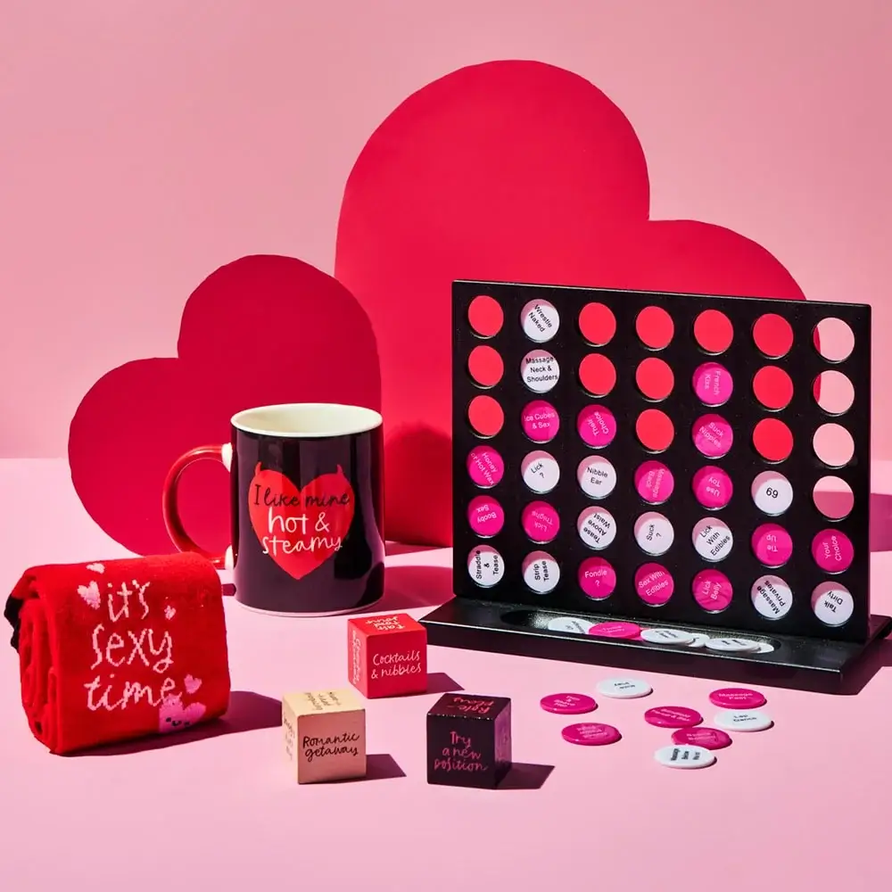 Valentine-themed gift set featuring a black mug with red text, a red towel with white text, a black and pink Connect Four-style game, and decorative heart-shaped props in the background.