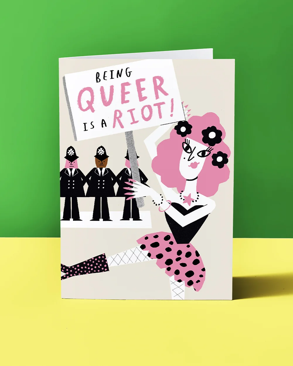 A Pride greeting card featuring an illustrated character holding a protest sign reading ‘Being Queer is a Riot!’, with stylised figures in uniform in the background, displayed against a green backdrop with a yellow surface.