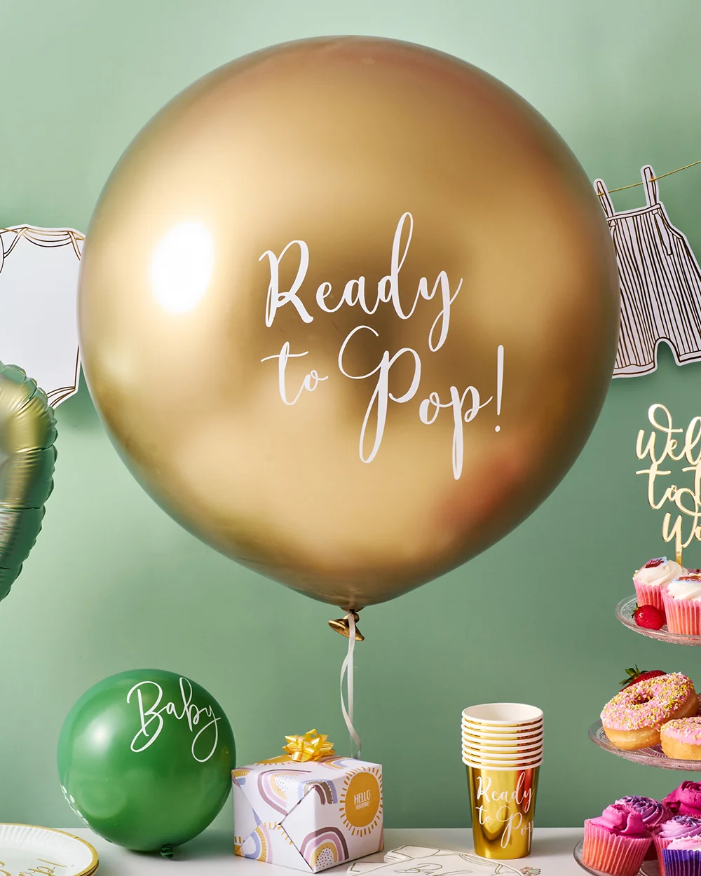A baby shower display featuring a large gold balloon printed with the words ‘Ready to Pop!’ against a sage green backdrop. The setup includes baby-clothes bunting, a green balloon printed with ‘Baby,’ a rainbow-patterned gift box, gold party cups, baby shower tableware, and a tiered stand filled with pink cupcakes and sprinkled doughnuts, creating a modern baby shower celebration theme. A baby shower display featuring a large gold balloon printed with the words ‘Ready to Pop!’ against a sage green backdrop. The setup includes baby-clothes bunting, a green balloon printed with ‘Baby,’ a rainbow-patterned gift box, gold party cups, baby shower tableware, and a tiered stand filled with pink cupcakes and sprinkled doughnuts, creating a modern baby shower celebration theme.