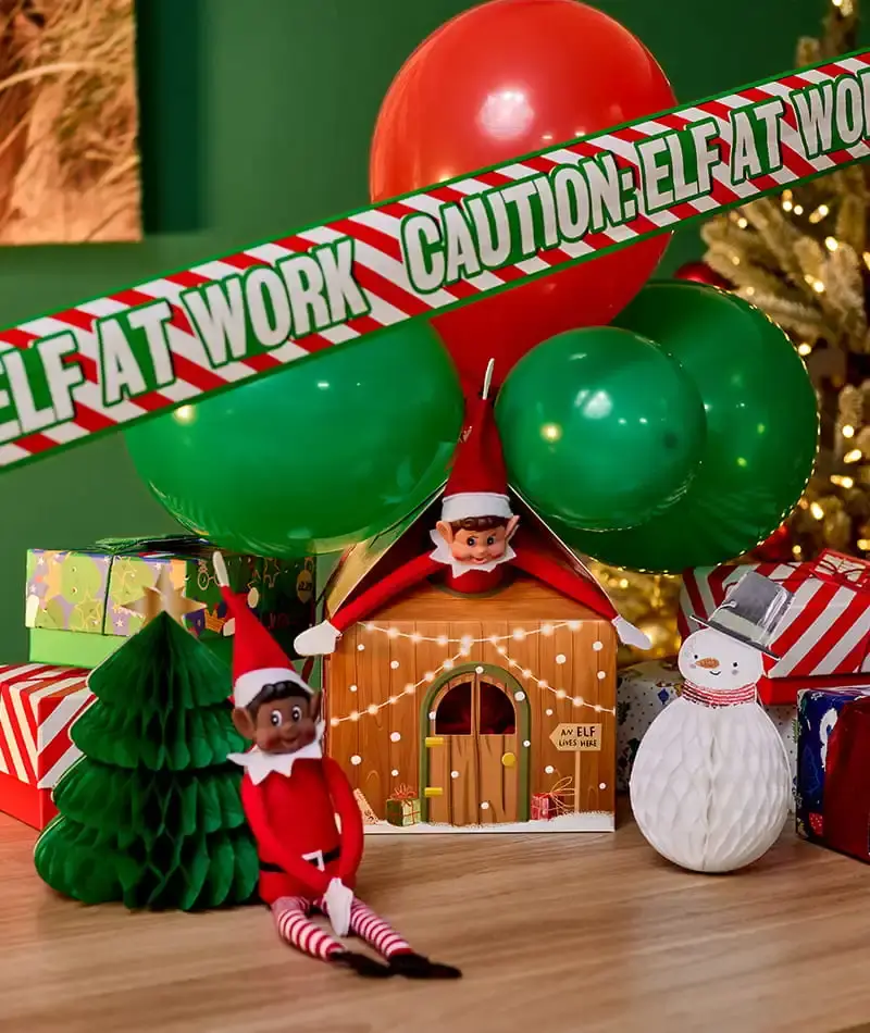 Elf on the Shelf Christmas display featuring festive balloons, an elf caution tape banner, gift boxes, honeycomb decorations and elf characters around a wooden elf house.