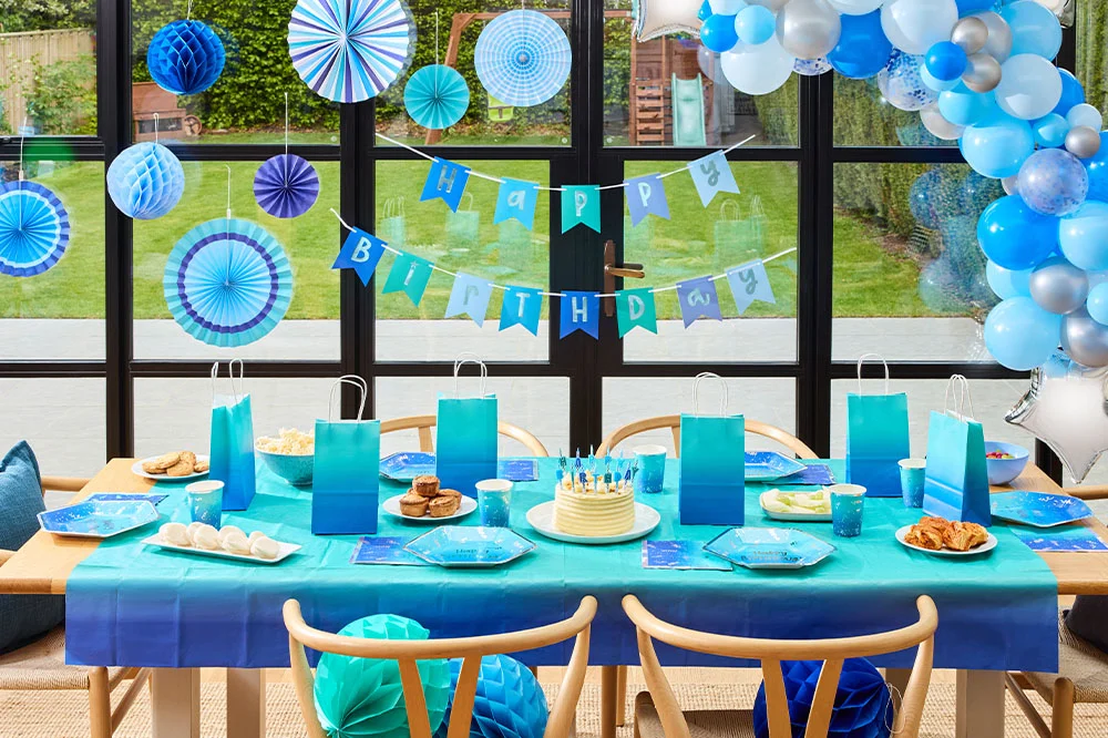 A blue-themed birthday party display featuring a table set with ombré blue party bags, plates, cups and napkins, centred around a birthday cake with blue candles. Behind the table, blue paper fan decorations, hanging honeycomb ornaments, a ‘Happy Birthday’ bunting banner and a blue, white and silver balloon garland create a coordinated indoor party backdrop. A blue-themed birthday party display featuring a table set with ombré blue party bags, plates, cups and napkins, centred around a birthday cake with blue candles. Behind the table, blue paper fan decorations, hanging honeycomb ornaments, a ‘Happy Birthday’ bunting banner and a blue, white and silver balloon garland create a coordinated indoor party backdrop.