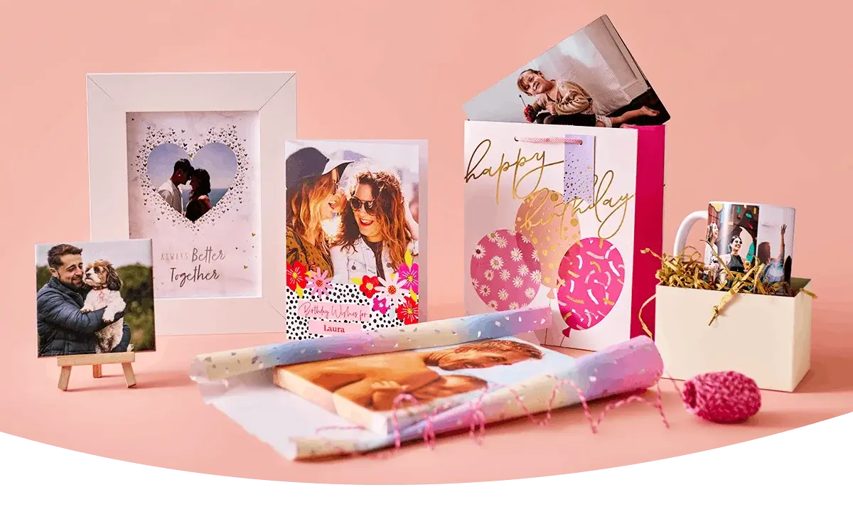 A display of personalised photo gifts on a peach background, including a photo frame, greeting cards, a birthday gift bag, a photo mug, and wrapped photo prints.