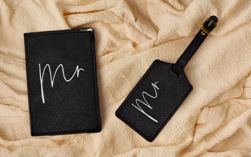 A black passport holder and matching luggage tag set, both printed with ‘Mr’ in white script, placed on soft beige fabric. A black passport holder and matching luggage tag set, both printed with ‘Mr’ in white script, placed on soft beige fabric.