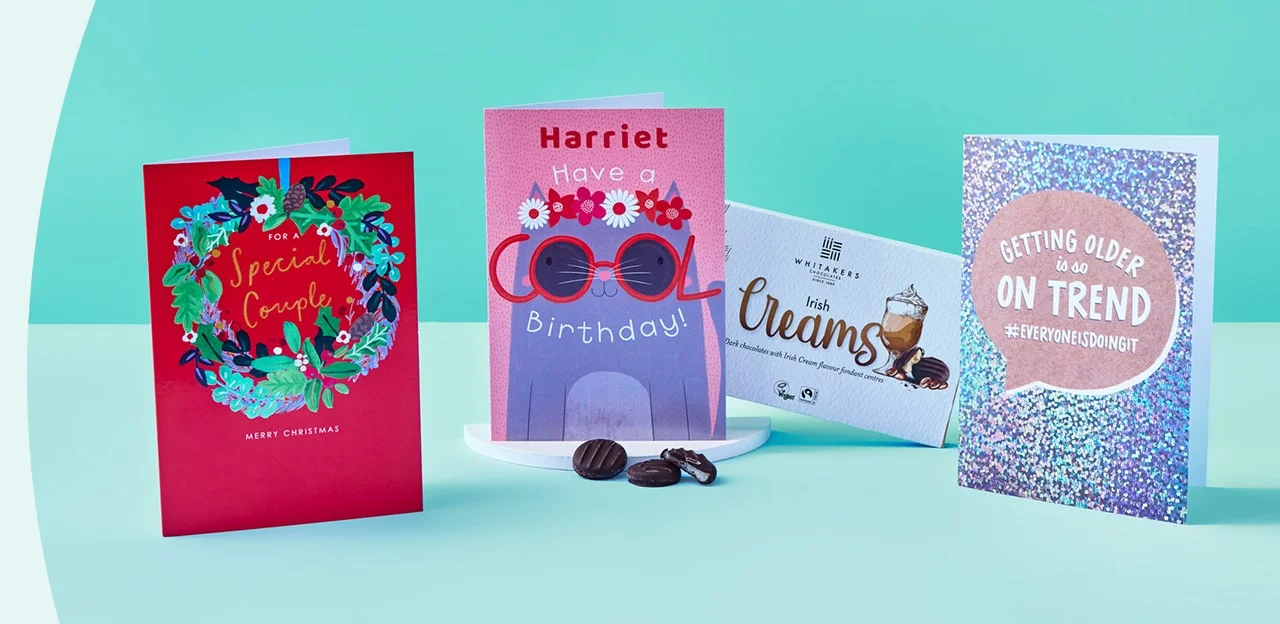 Four greeting cards and a box of chocolates arranged on a light blue surface with a teal background, including a red Christmas card with a wreath, a pink birthday card with sunglasses and flowers, a box of Whitakers Irish Creams, and a silver glittery card with a humorous message about getting older.