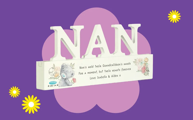 A decorative mantel sign featuring large white letters spelling ‘NAN,’ with a floral illustrated base and a printed sentimental message, shown against a purple background with a pink flower shape and yellow daisies.