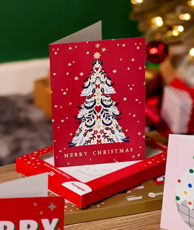 A boxed Christmas card set featuring a white card with a decorated Christmas tree and gold ‘Merry Christmas’ lettering, displayed on festive packaging with Christmas decorations in the background.