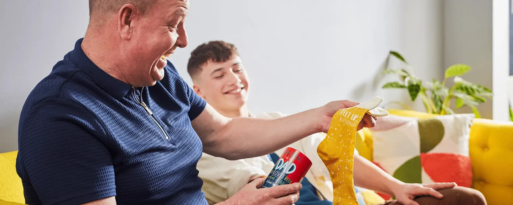 Two people seated on a sofa while one holds a small gift tin and the other receives a pair of yellow socks, with cushions and houseplants visible in the background. Two people seated on a sofa while one holds a small gift tin and the other receives a pair of yellow socks, with cushions and houseplants visible in the background.