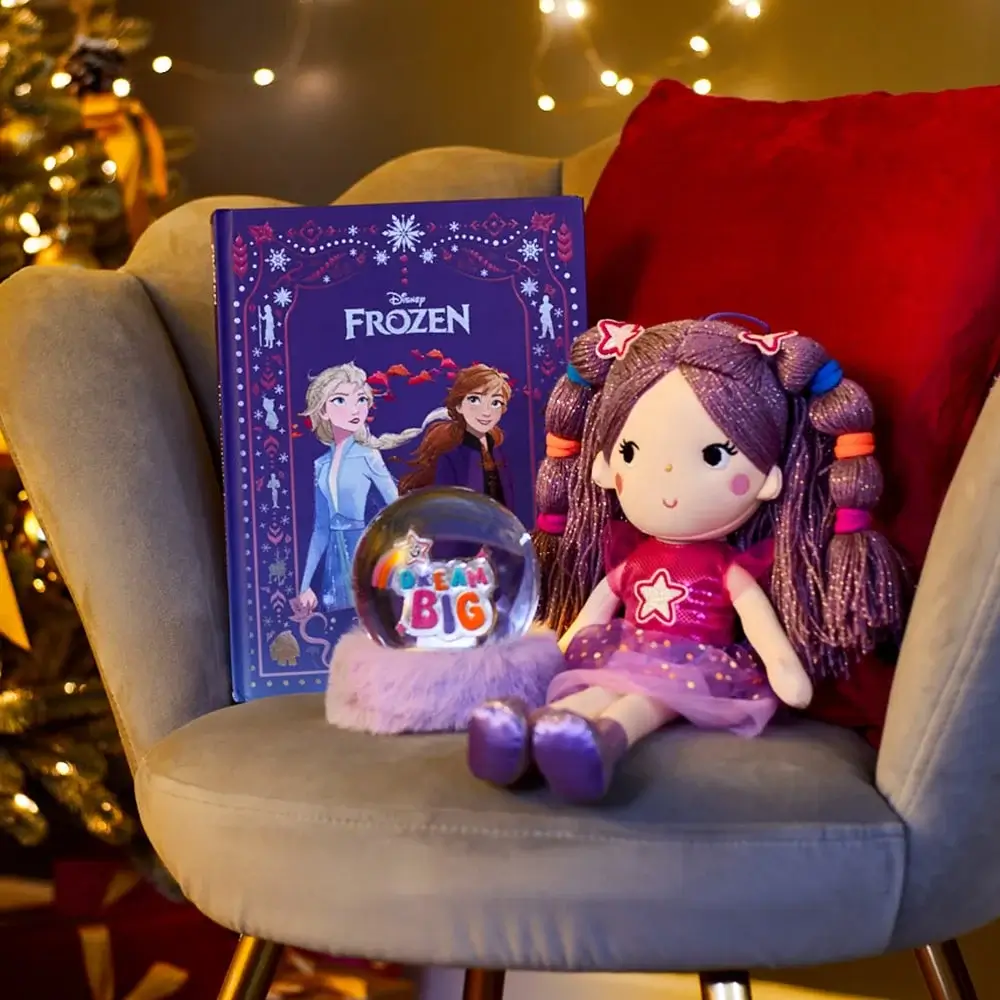 Purple Frozen-themed book, a plush doll with purple hair and pink dress, and a snow globe with the words Dream Big placed on a gray chair with a red cushion in a festive setting.
