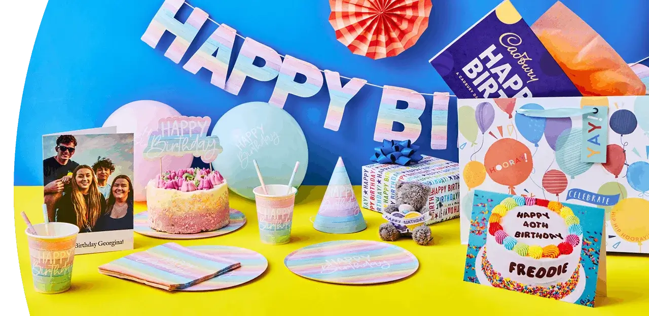 Colourful birthday party display with pastel tableware, a decorated cake, wrapped gifts, party hats, and greeting cards arranged against a bright backdrop.