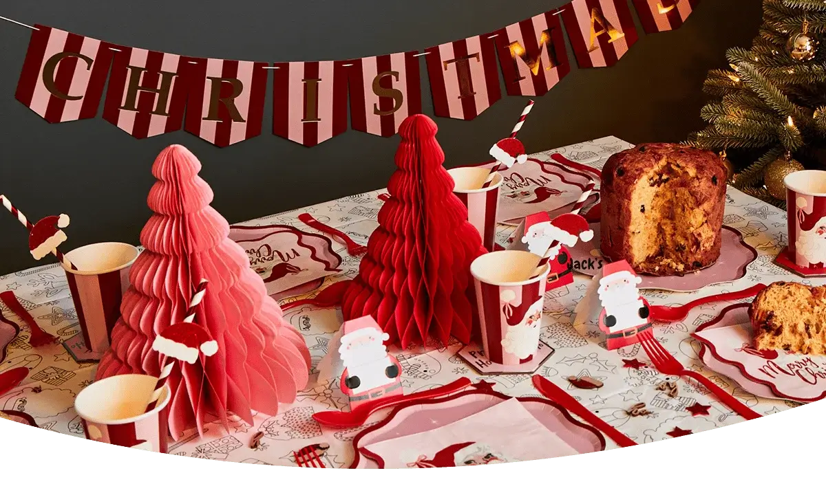 Christmas-themed party table with red and pink paper trees, Santa decorations, striped cups, festive plates, and a loaf of panettone under a red and white Christmas banner. Christmas-themed party table with red and pink paper trees, Santa decorations, striped cups, festive plates, and a loaf of panettone under a red and white Christmas banner.