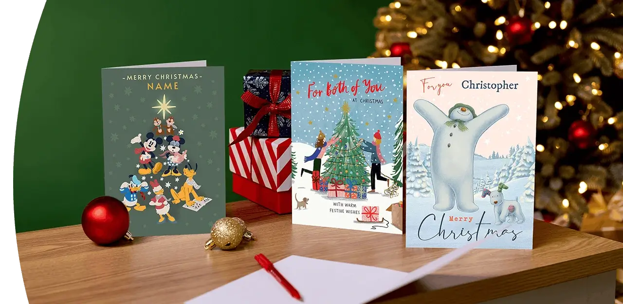 Selection of Christmas cards, including Disney, festive and personalised designs, displayed on a wooden table with wrapped presents and a decorated Christmas tree in the background.