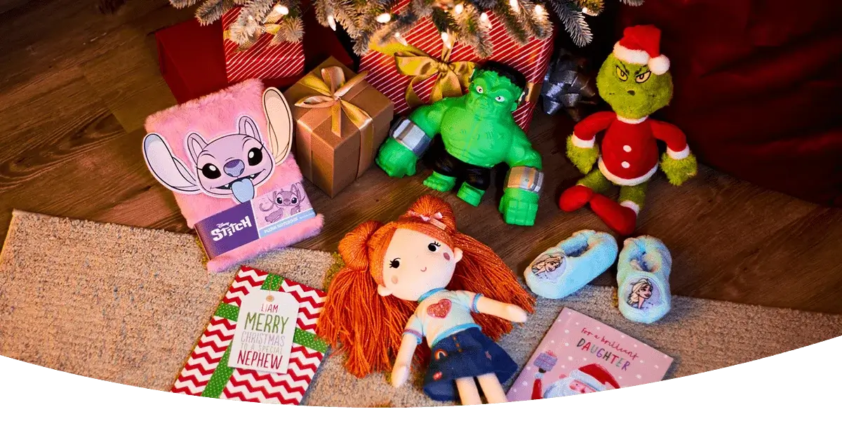 Christmas gifts arranged on a wooden floor, including a pink Stitch notebook, wrapped presents, a green Hulk figure, a Grinch plush toy in a Santa outfit, a doll with orange yarn hair, blue Frozen slippers, and two festive greeting cards.
