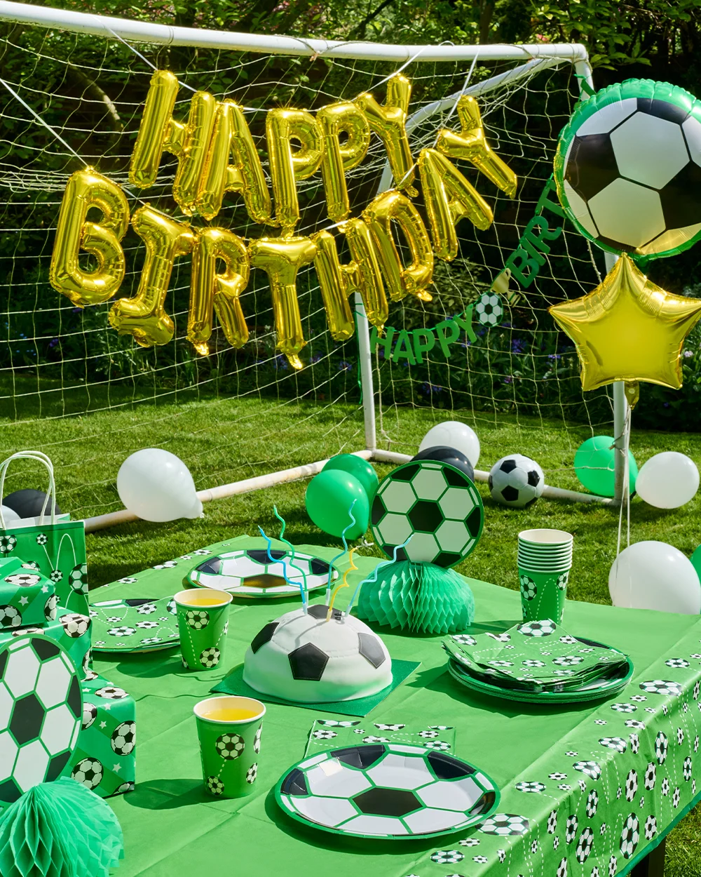 A football-themed birthday party table set up outdoors in front of a goal net, decorated with green football-print tableware including plates, cups, napkins and a football-patterned tablecloth. The display features a football-shaped birthday cake, football honeycomb centrepieces, themed gift bags and party boxes, with gold foil ‘Happy Birthday’ balloons hanging across the goal. Football and star-shaped foil balloons, along with green, white and black balloons, complete the celebration scene.
