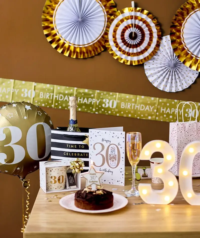 30th birthday celebration table with a birthday cake, champagne, gift bags, a 30 balloon, illuminated number decorations, and gold party décor.