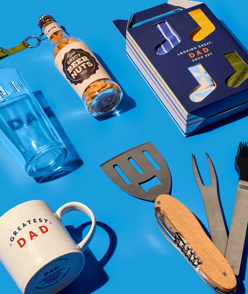 Father’s Day gift set with greatest dad mug, spicy beer nuts, socks in a gift box, and BBQ tools arranged on a blue background Father’s Day gift set with greatest dad mug, spicy beer nuts, socks in a gift box, and BBQ tools arranged on a blue background