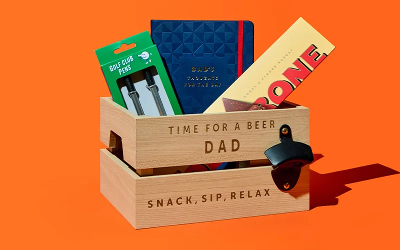 A wooden gift crate engraved with the text ‘Time for a Beer Dad’ and ‘Snack, Sip, Relax,’ filled with Father’s Day items including a pack of golf club pens, a blue notebook that reads ‘Dad’s Notes for the Day,’ and a Toblerone chocolate bar, with a bottle opener attached to the front, displayed against a bright orange background. A wooden gift crate engraved with the text ‘Time for a Beer Dad’ and ‘Snack, Sip, Relax,’ filled with Father’s Day items including a pack of golf club pens, a blue notebook that reads ‘Dad’s Notes for the Day,’ and a Toblerone chocolate bar, with a bottle opener attached to the front, displayed against a bright orange background.