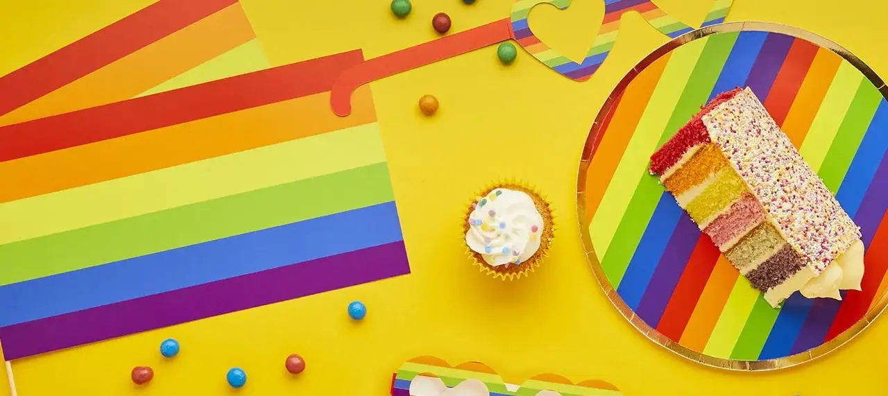 'A colorful scene with a slice of rainbow layer cake on a rainbow-striped plate, a cupcake with white frosting and sprinkles, rainbow-striped paper decorations, and scattered colorful candies on a bright yellow background.'