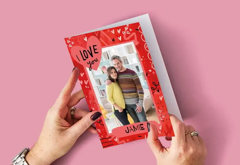 Hands holding a red love‑themed greeting card featuring a couple’s photo and the text I love you and Jamie.