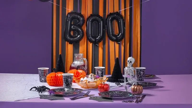 Halloween-themed table setting with black and orange decorations, including 'BOO' balloons, spiderweb-patterned tableware, a red drink pitcher, pumpkins, a skeleton figurine, and spooky accents.