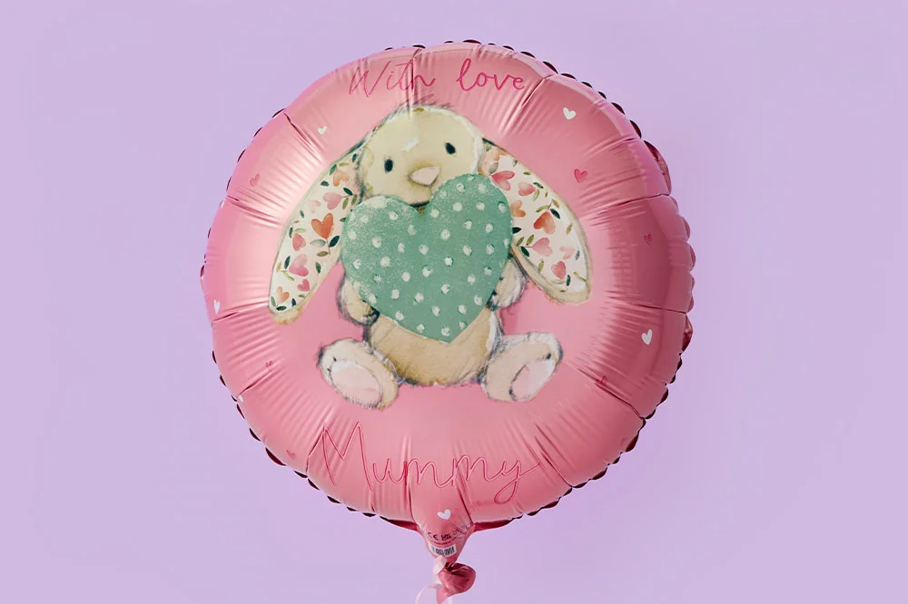 A round pink foil balloon featuring an illustration of a bunny holding a green heart with patterned ears, and the text ‘With love Mummy’ on a pastel purple background.