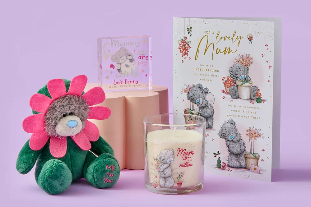 A Tatty Teddy flower‑costumed plush toy, a scented candle, a decorative acrylic block, and a floral Mother’s Day greeting card arranged together on a pastel purple background.