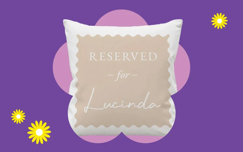 A white cushion with a beige panel reading ‘Reserved for Lucinda,’ displayed against a purple background with a pink flower shape and small yellow daisies.