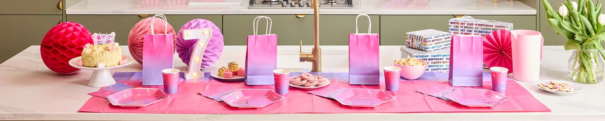 A pink and purple ombré birthday party table arranged in a modern kitchen, featuring matching gift bags, plates, cups and napkins displayed around a birthday cake topped with a ‘Happy Birthday’ decoration. Light-up number 7 lights, wrapped presents, paper honeycomb decorations, sweet treats and pink party accessories complete the coordinated celebration display.”