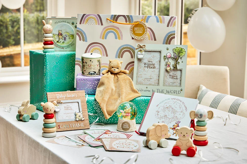 A collection of baby gifts and keepsakes arranged on wrapped presents, including Winnie the Pooh photo frames, a soft comforter toy, children's books, greeting cards, wooden animal toys, stacking toys and a decorative snow globe. The display is styled as a literary-themed baby shower or new baby gift arrangement with white balloons in the background. A collection of baby gifts and keepsakes arranged on wrapped presents, including Winnie the Pooh photo frames, a soft comforter toy, children's books, greeting cards, wooden animal toys, stacking toys and a decorative snow globe. The display is styled as a literary-themed baby shower or new baby gift arrangement with white balloons in the background.