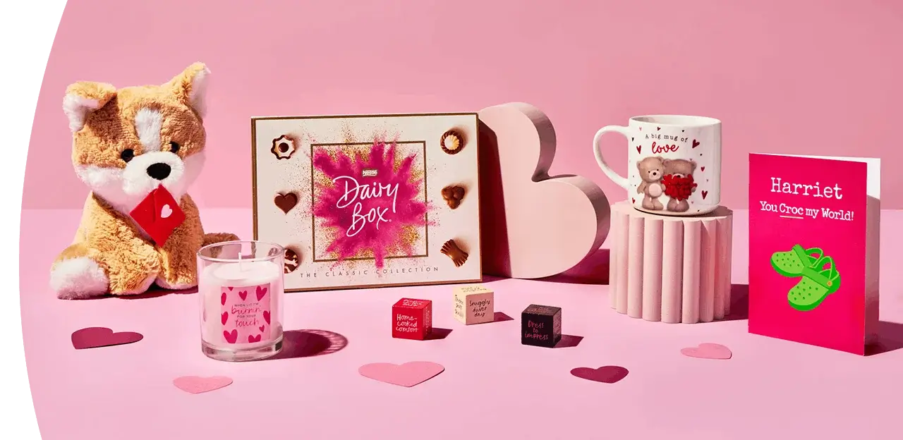 A pink Valentine’s‑themed display featuring a plush dog, a box of chocolates, a heart‑decorated candle, small gift cubes, a cute illustrated mug, and a bright greeting card arranged with scattered paper hearts.