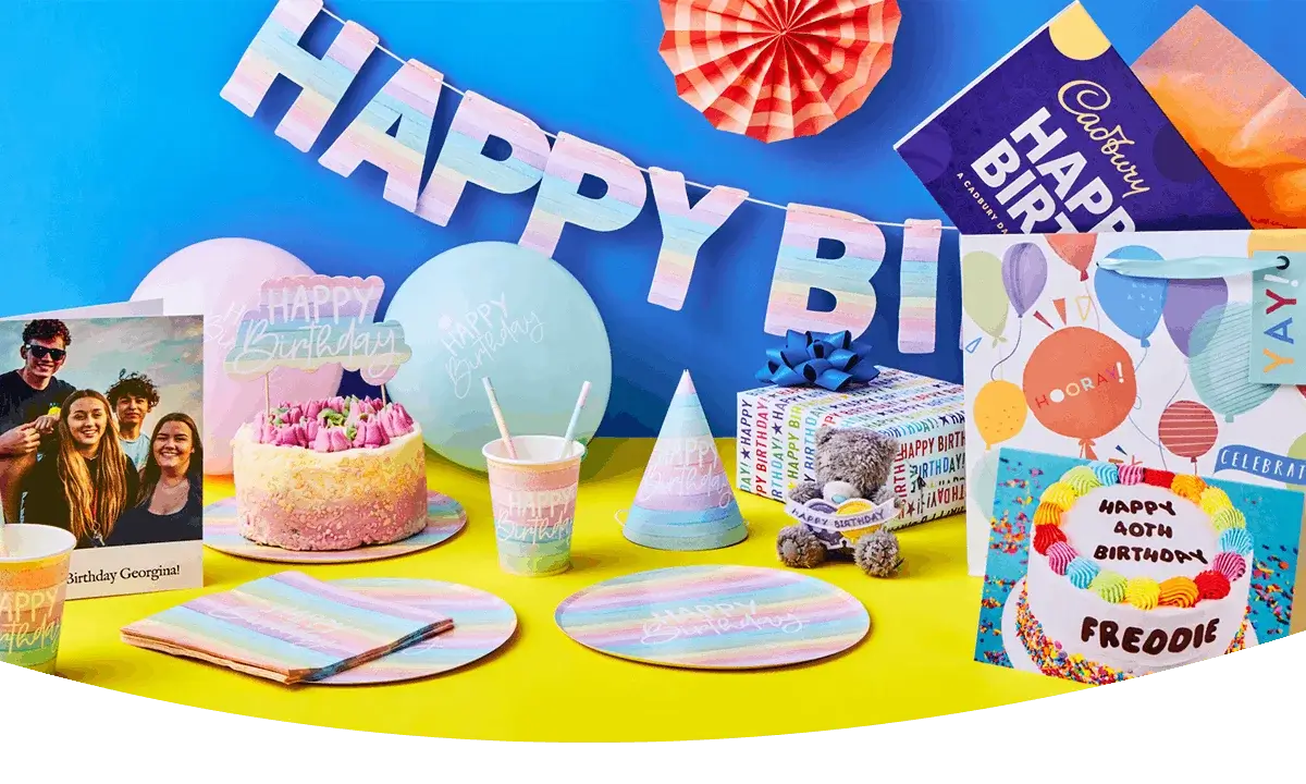 Colourful birthday party display with pastel tableware, a decorated cake, wrapped gifts, party hats, and greeting cards arranged against a bright backdrop.
