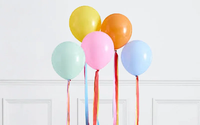 A cluster of five helium balloons in pastel yellow, orange, pink, blue and mint green, each decorated with long multicoloured ribbon tails, floating against a white panelled wall. A cluster of five helium balloons in pastel yellow, orange, pink, blue and mint green, each decorated with long multicoloured ribbon tails, floating against a white panelled wall.