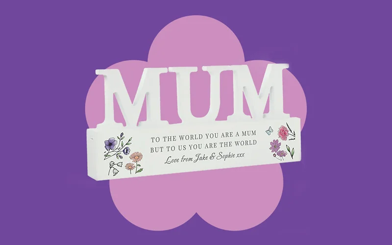 A white wooden ornament spelling ‘MUM,’ decorated with small floral illustrations and a heartfelt message on a purple background.