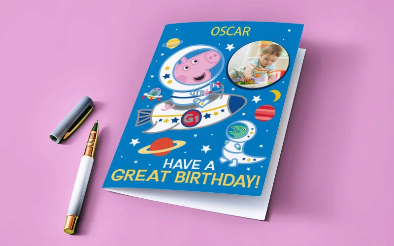 A personalised Peppa Pig birthday card with a space theme, showing Peppa Pig in a rocket, planets and stars, a circular photo window, and the text ‘Have a Great Birthday!’ with the name ‘Oscar’ printed at the top, displayed on a pink background with a pen beside it.