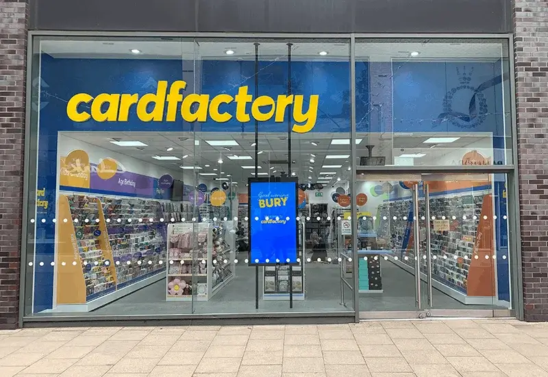 Card Factory store entrance with greeting card displays, bright blue branding, and aisles of cards visible through the glass frontage.