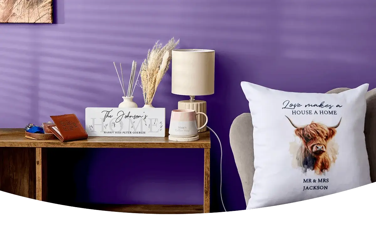 A cosy home display featuring personalised wall art, a wooden clock, a table lamp, framed prints, decorative accessories and a cushion with a highland cow design arranged against a dark blue wall.