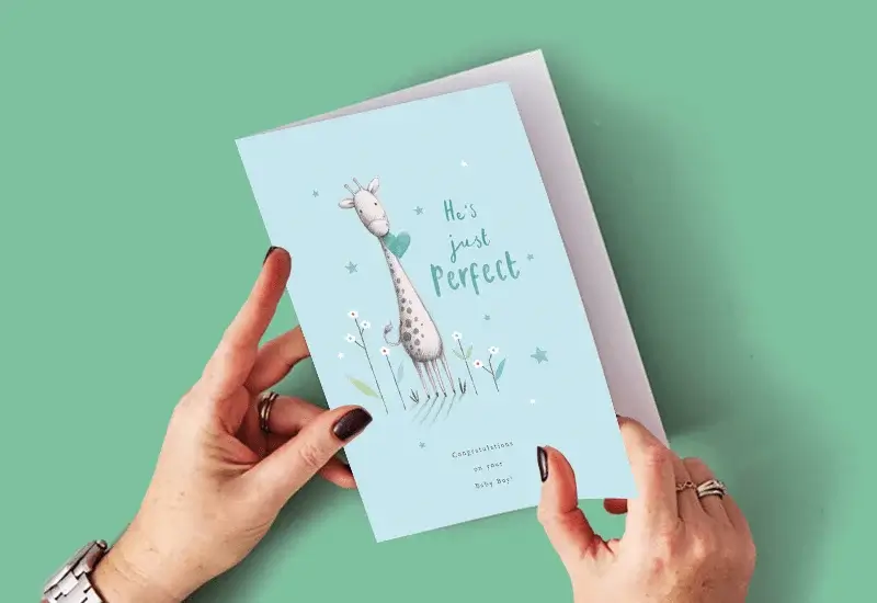 A person holding a light blue new baby card with a giraffe illustration, stars, flowers, and the text He's just perfect and Congratulations on your Baby Boy against a green background.