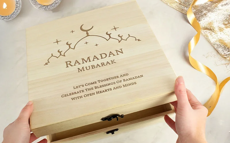 A light wooden keepsake box being opened, engraved with a crescent moon design, the words ‘Ramadan Mubarak,’ and a message about celebrating blessings.