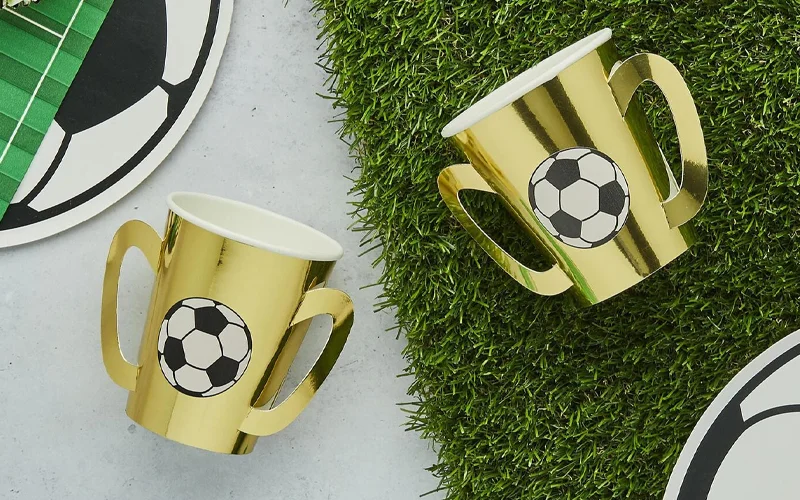 Two gold trophy-shaped party cups decorated with football graphics, displayed beside football-themed tableware and artificial grass in a football party setting. Two gold trophy-shaped party cups decorated with football graphics, displayed beside football-themed tableware and artificial grass in a football party setting.