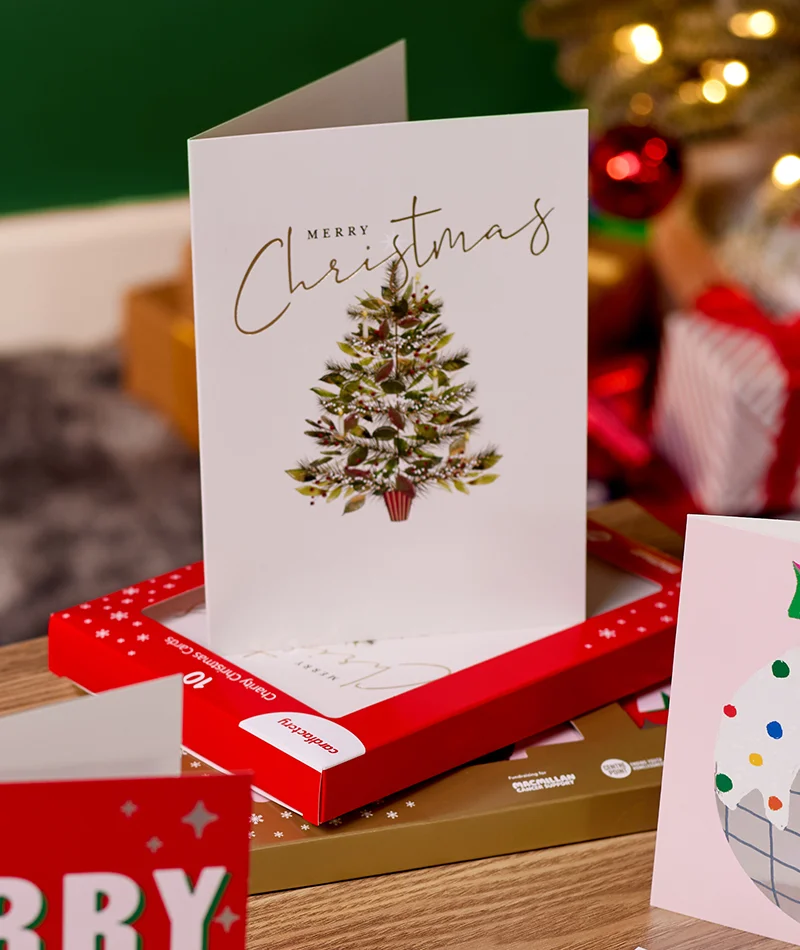 A boxed Christmas card set featuring a white card with a decorated Christmas tree and gold ‘Merry Christmas’ lettering, displayed on festive packaging with Christmas decorations in the background. A boxed Christmas card set featuring a white card with a decorated Christmas tree and gold ‘Merry Christmas’ lettering, displayed on festive packaging with Christmas decorations in the background.