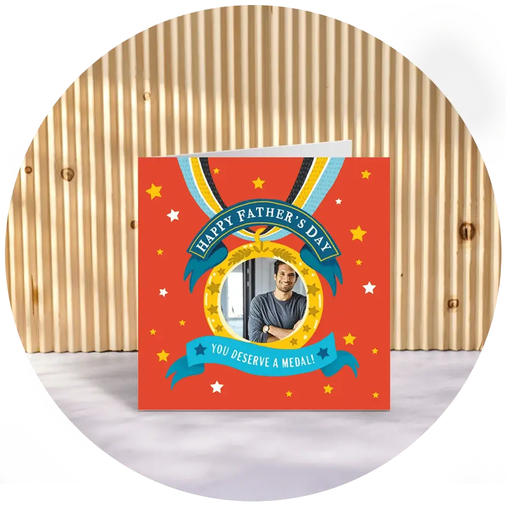 A Father’s Day card with a red background featuring a medal illustration and a circular photo window, with the text ‘Happy Father’s Day – You Deserve a Medal,’ displayed upright against a wooden slatted backdrop. A Father’s Day card with a red background featuring a medal illustration and a circular photo window, with the text ‘Happy Father’s Day – You Deserve a Medal,’ displayed upright against a wooden slatted backdrop.