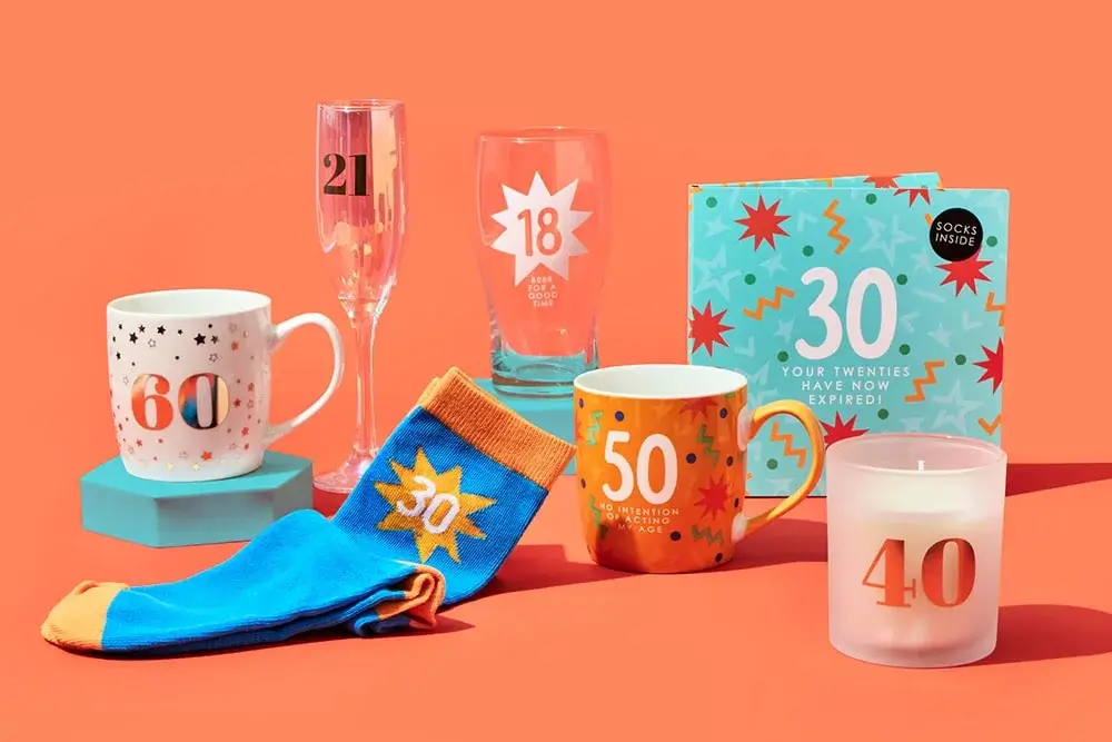 'The image shows a collection of birthday-themed items against an orange background. There is a white mug with the number 60 in red and orange, a champagne flute with the number 21 in black, a beer glass with the number 18 and text below it, a pair of blue and orange socks with the number 30, an orange mug with the number 50 and text below it, a white candle with the number 40 in red-orange, and a gift box with the number 30 and text 'YOUR TWENTIES HAVE NOW EXPIRED' along with 'SOCKS INSIDE' 'The image shows a collection of birthday-themed items against an orange background. There is a white mug with the number 60 in red and orange, a champagne flute with the number 21 in black, a beer glass with the number 18 and text below it, a pair of blue and orange socks with the number 30, an orange mug with the number 50 and text below it, a white candle with the number 40 in red-orange, and a gift box with the number 30 and text 'YOUR TWENTIES HAVE NOW EXPIRED' along with 'SOCKS INSIDE'