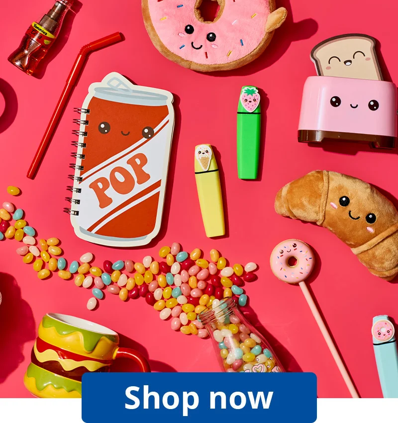 A bright flat‑lay of novelty gifts and stationery arranged on a pink background, including a croissant plush, toaster-shaped toy, donut plush, sweets, colourful pens, a spiral notebook with a smiling soda cup design, and a ‘Shop now’ button at the bottom.