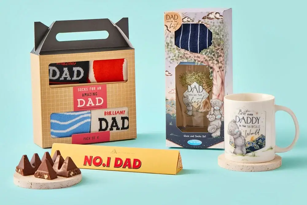 The image shows a collection of Father's Day gifts, including a box of socks labeled 'Socks for an amazing Dad,' a gift set with socks and a mug featuring the phrase 'Best Daddy in the whole wide world,' and a Toblerone chocolate bar with the text 'No.1 Dad.' The image shows a collection of Father's Day gifts, including a box of socks labeled 'Socks for an amazing Dad,' a gift set with socks and a mug featuring the phrase 'Best Daddy in the whole wide world,' and a Toblerone chocolate bar with the text 'No.1 Dad.'