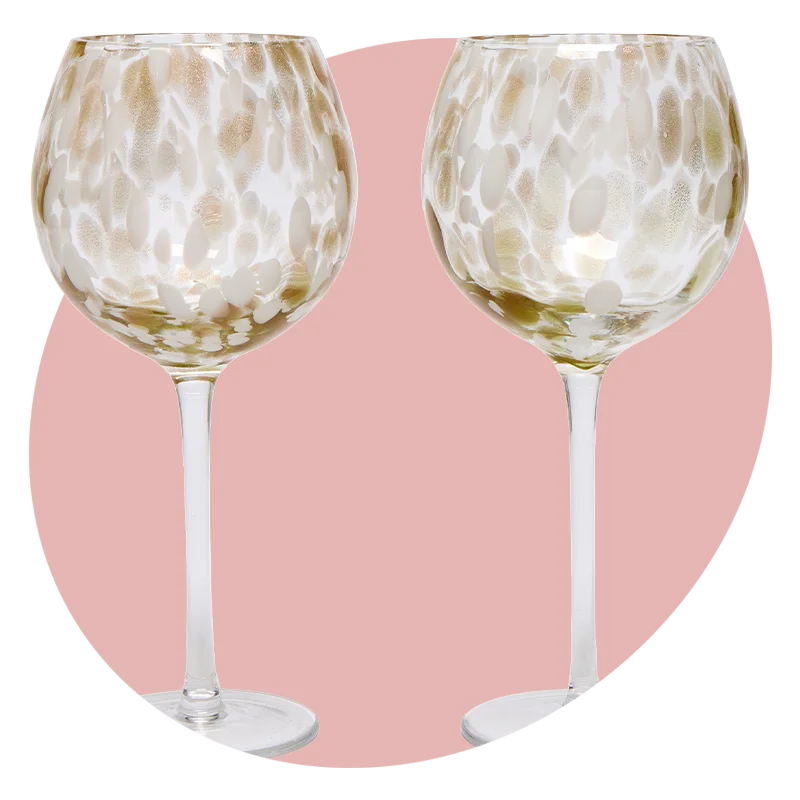 A set of two stemmed wine glasses featuring a gold and white abstract pattern, displayed against a soft pink circular background. A set of two stemmed wine glasses featuring a gold and white abstract pattern, displayed against a soft pink circular background.