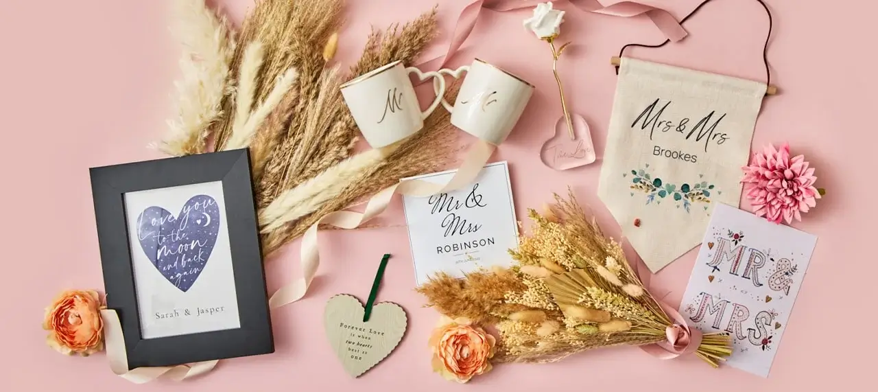 A wedding-themed flat lay on a pink background featuring items like two mugs labeled 'Mr' and 'Mrs', a framed picture with the text 'Love you to the moon and back - Sarah & Jasper', a heart-shaped wooden ornament reading 'Forever Love - Always & Forever', a banner with 'Mrs & Mrs Brookes', a bouquet of dried flowers, and cards that say 'Mr. & Mrs. ROBINSON' and 'MR. & MRS.'. Pink flowers accent the arrangement.